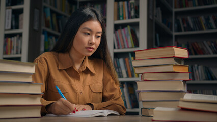 Smart focused asian woman girl student doing homework at desk with books in university library serious female schoolgirl writing notes preparing for college exam write scientific historical project