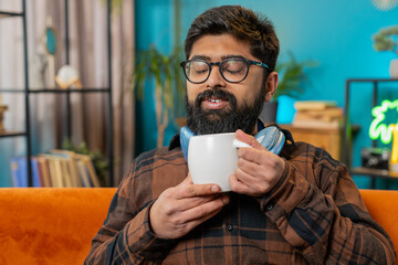 Smiling happy Indian young man drinking a cup of warm coffee or herbal tea sitting at home living room sofa in morning. Hispanic guy enjoying comfortable relaxing alone. Relaxing, taking a break