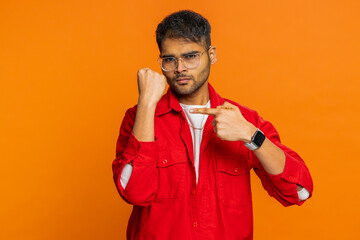 Aggressive angry Indian young man trying to fight at camera, shaking fist, boxing with expression, punishment, threaten, bullying, abuse, mad fury. Arabian guy isolated on orange studio background