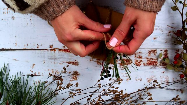 Close-up of hands carefully tying a twine bow on a gift, adding natural pine and berry accents. Rustic, festive wrapping scene perfect for holiday promotions, crafts, and seasonal content.