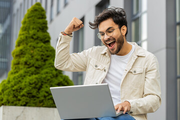 Overjoyed happy surprised Indian man working on laptop scream in delight raise hands in triumph...