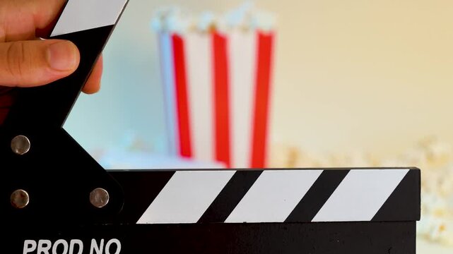 Male hands closing a black and white wooden clapperboard with a cardboard bucket full of popcorn and blue and red 3D glasses next to it, concept of film production and food