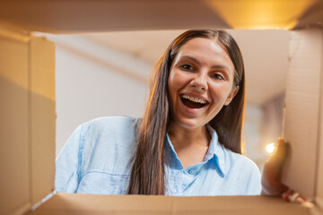 Happy woman unpacking delivery parcel at home. Smiling satisfied young girl shopper online shop customer opening cardboard box receiving purchase gift by fast postal shipping. View inside from the box