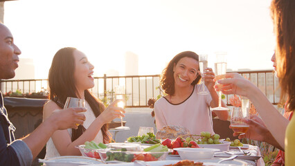 Friends Making Toast Gathered On Rooftop Terrace For Meal With City Skyline In Background 