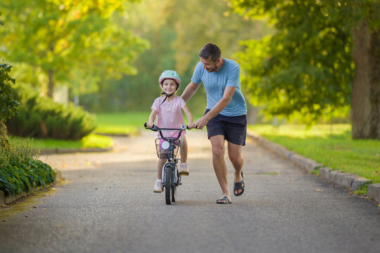 Young adult father teaching happy beautiful little girl to ride on first bike on sidewalk at city park. Learning to keep balance. Warm summer day. Cute 5 years old toddler. Front view.