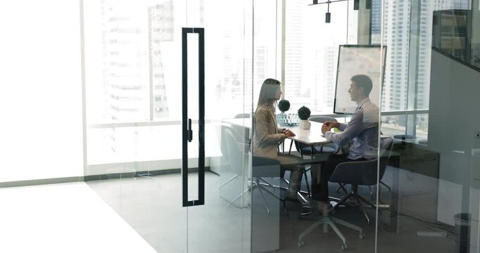 Two business partners sit across from each other in office with large window view, negotiate behind closed glass wall in modern skyscraper conference room. Collaboration and professional communication