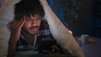 Gadget internet social media addict guy Latino tired man Arabian confused worried puzzled Indian male at night evening dark home under blanket cover with duvet scrolling mobile phone upset frustrated