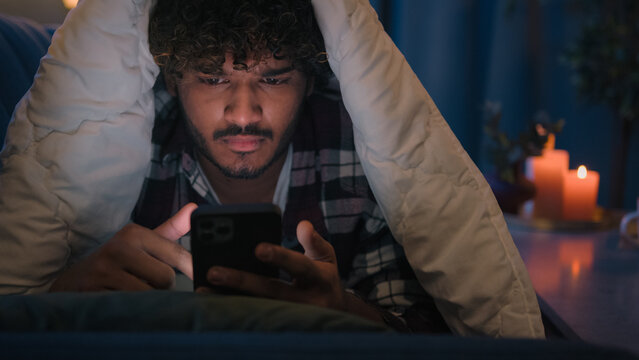 Gadget internet social media addict guy Latino tired man Arabian confused worried puzzled Indian male at night evening dark home under blanket cover with duvet scrolling mobile phone upset frustrated