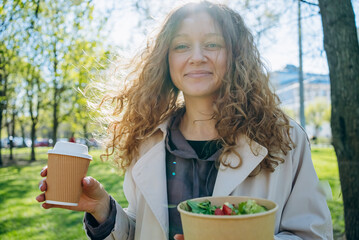 Young woman with curly hair is holding a takeaway coffee cup and salad bowl while enjoying her...