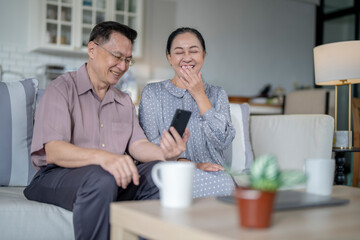 An elderly couple sits together in their cozy living room, smiling and enjoying a moment as they look at something on a smartphone. Their warmth and affection capture a peaceful home atmosphere.