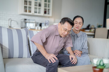 An elderly couple sits in their living room. The man holds his knee, appearing to be in discomfort, while his partner offers support. The image conveys care, concern, and the bond between the couple.