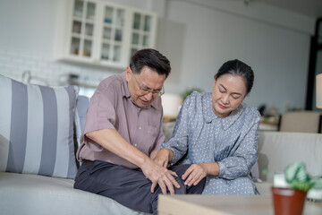 An elderly couple sits in their living room. The man holds his knee, appearing to be in discomfort, while his partner offers support. The image conveys care, concern, and the bond between the couple.