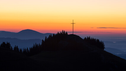 View of the serene summit with a cross under a breathtaking sunrise sky, Brandkogel, Voitsberg, Styria, Austria.