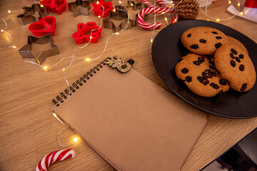 Top down view of cozy Christmas setup, plate of chocolate chip cookies, festive candy canes, cookie cutters, spiral notepad with reindeer clip. Wooden table, recipe themes or festive planning