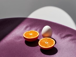 An egg and an orange cut in half on a purple surface with a light reflection on the top of the egg.
