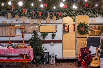 Cozy outdoor Christmas setting featuring a decorated camper, picnic table, string lights, and holiday-themed elements like wreaths, stockings, and a guitar for seasonal warmth.