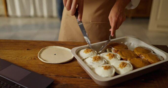 A pastry chef girl wearing a shirt and a running apron takes out a Cinnabon cake from a baking sheet using a knife and a pastry spatula in a bakery