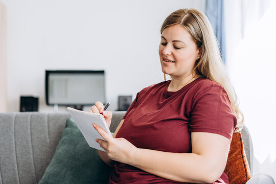 Smiling plus size woman making notes sitting on sofa at home