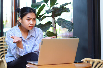 confused asian woman sitting in cafe working on laptop with raising palms show i do not know...