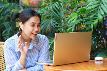 excited asian woman looking at laptop having video chat online meeting with raised palms say hi gesture while sitting in cafe outdoor with green plant nature background wearing striped blue shirt