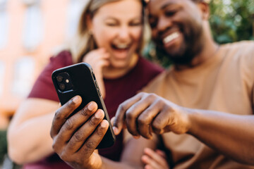 Fototapeta premium Happy multiethnic couple looking at smartphone screen sitting outside