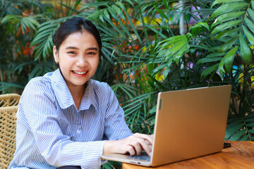 excited asian woman sitting in outdoor cafe with green plant interior using laptop working from anywhere, talking on computer have online meeting video chat