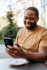 Happy African American man drinking coffee using smartphone at cafe outdoors