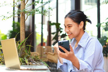 excited asian woman working on laptop and holding smartphone working from outdoor cafe have online meeting with victory gesture