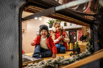 Fototapeta premium Smiling boy in a red jacket and sister in earmuffs pose in a cozy camper kitchen, decorated with Christmas lights and holiday accents. Warm family bonding during the holidays.