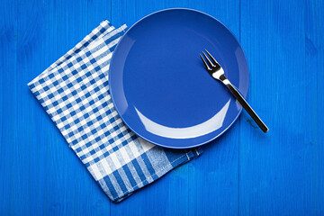 empty empty blue plate with fork and checkered kitchen towel on wooden blue table, top view