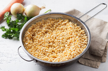 Yellow split soaked peas in a metal colander, onion, carrot and parsley, stage of cooking, on a light gray background