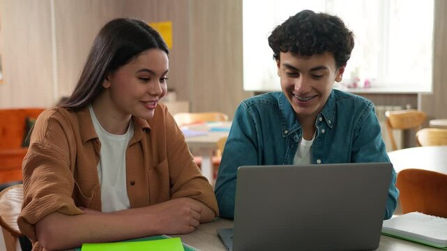 Two Caucasian students pupils friends man woman girl guy boy female male studying together learning homework school project e-learning laptop computer talking high five cooperation teamwork education