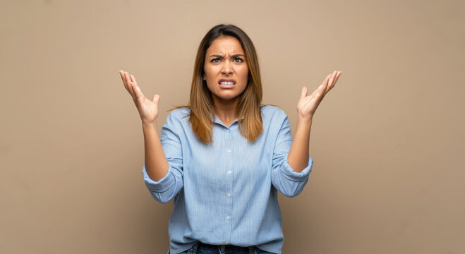 Portrait of a woman standing arms raised with exasperated and angry expression on beige background.