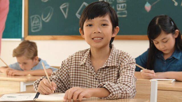 Asian smart boy smiling to camera while student writing answer in answer sheet. Multicultural student doing classwork or test while caucasian teacher checking student homework at classroom. Pedagogy.