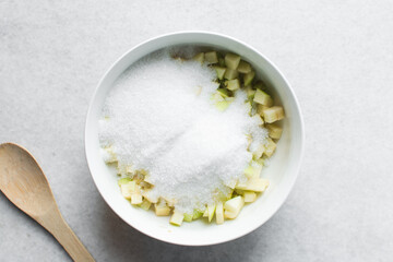 Overhead view of diced green apples and sugar being mixed together in a white bowl, process of making apple cheong or apple syrup