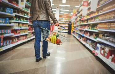 Woman, basket and legs of customer in supermarket, goods discount and shelves for deal. Female person, sale and shop for options on offer, retail purchase and browsing for grocery choice in aisle