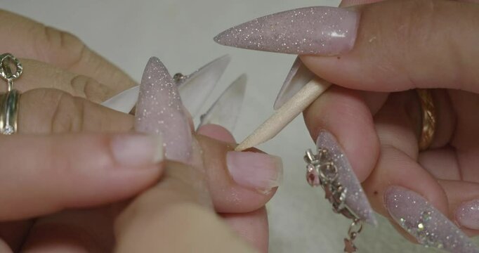 A manicurist skillfully removes cuticles from acrylic nails with a wooden tool in a beauty salon. A close-up of the detailed manicure process in a beauty salon is shown. Part of a series.