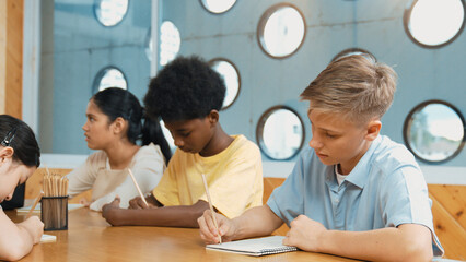 Fototapeta premium Smart boy listening teacher explain while lecturing and think about idea. Diverse student study or learning together in classroom while writing idea or taking a note. Creative education. Edification.