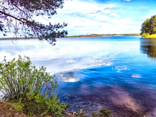 Calm lake reflects the sky, surrounded by greenery and distant hills under bright sunlight.