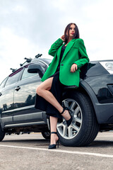 A girl in a green jacket poses near a black car in a parking lot.