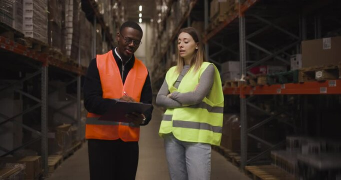 Warehouse workers discussing logistics with clipboard in storage facility wearing safety vests for organization and management in industrial environment