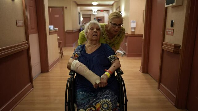 Family daughter visiting, pushing mother with broken arm in wheelchair in rehab facility.