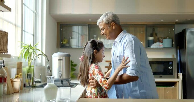 An Indian Asian father in his late forties playfully encourages his small daughter to drink milk while she sits happily on the kitchen platform, creating a joyful moment filled with love and laughter