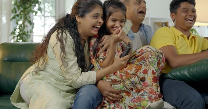 Indian Asian family of four, parents in their forties and two kids, sitting closely on a sofa, laughing out loud while watching television together, enjoying a fun-filled family evening at home