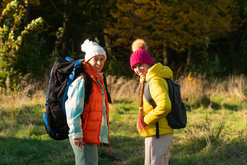  family walking in autumn forest with backpacks, boy in yellow jacket, girl in pink jacket, mother in orange sleeveless jacket, family in hat with pompoms, gen z