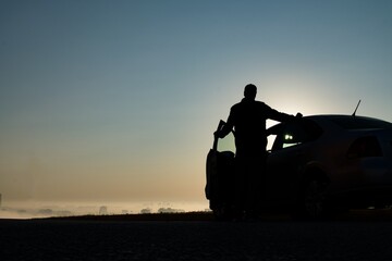 The silhouette of a man standing by an open car against the background of the city in the fog at dawn