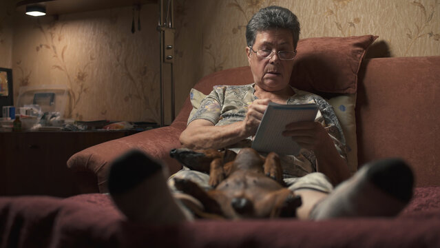 Elderly woman sitting on sofa and solving crossword puzzle, holding dachshund.