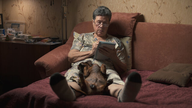 Elderly woman sitting on sofa and solving crossword puzzle, holding dachshund.