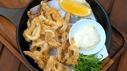 Crispy calamari rings served with lemon wedge, dip sauce and fresh arugula on wooden table.