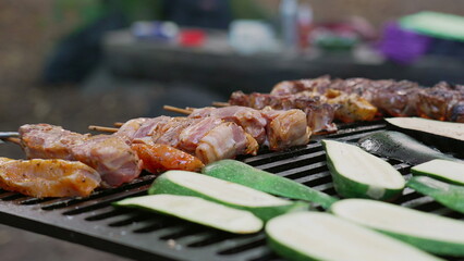 Close-up of skewers and vegetables being grilled over a flame, showing a variety of delicious food cooking outdoors, including zucchini and meat